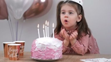 Little girl blows out candles on a birthday cake at home against a backdrop of balloons. Childs birthday. 4K.