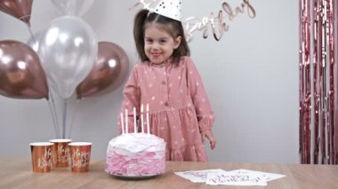 Cute little girl stands near a cake with candles in a festively decorated room. Child celebrates a birthday. 4K.