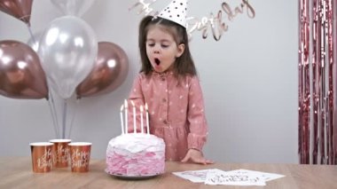 Little girl blows out candles on a birthday cake at home against a backdrop of balloons. Childs birthday. 4K.