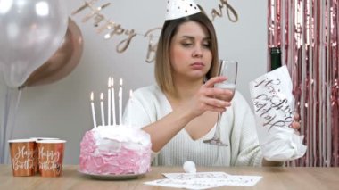 Beautiful young woman drinking wine alone. She sits against the backdrop of a festively decorated wall in front of a birthday cake and is sad. HD.