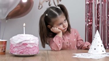 Üzgün küçük kız bir doğum günü pastasının yanındaki şenlik masasında oturuyor. 4K.