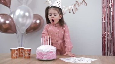 Little girl blows out candles on a birthday cake at home against a backdrop of balloons. Childs birthday. 4K.