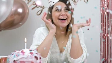 Beautiful young woman celebrates her birthday. Nicely decorated house, colorful confetti shower. Girl blows out a candle on a birthday cake. HD.
