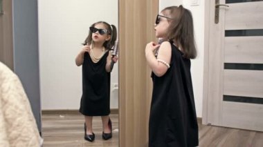 Annesinin ayakkabıları, güneş gözlükleri ve elinde saç fırçası olan küçük bir kız aynanın önünde bir şarkıcıyı resmediyor. HD.