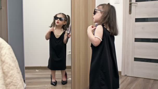 母の靴にサングラスをかけ 手にヘアブラシをかけた少女が鏡の前で歌手を描いている — ストック動画