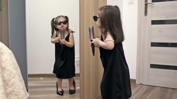 母の靴にサングラスをかけ 手にヘアブラシをかけた少女が鏡の前で歌手を描いている — ストック動画