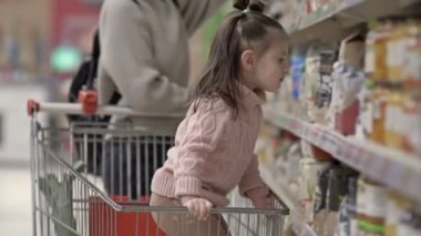 Markette ürünleri olan, rafların yanında çocuğu olan genç bir anne. Küçük kız kendi başına raftan bir paket almaya çalışıyor. 4K.