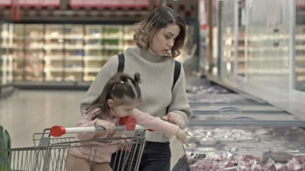 彼女の小さな娘と一緒に若い女性はスーパーマーケットで肉とショーケース 子供は食料品のカートに座って お母さんが選択をするのを助けます — ストック動画