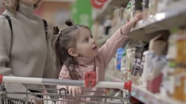 Markette ürünleri olan, rafların yanında çocuğu olan genç bir anne. Küçük kız raftan kendi başına bir paket aldı. 4K.