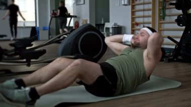 Kilolu bir adam spor salonunda paspasın üzerine uzanmış karın kaslarını özenle itiyor. 4K.