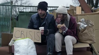 İki kirli ve kötü giyimli evsiz insan, bir adam ve bir kadın, ellerinde bir CRISIS posteriyle bir çöp yığınının yanında oturmuş bir şeyler yiyorlar. 4K.