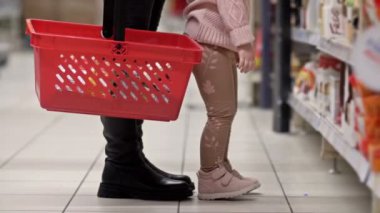 Markette rafın yanında elinde kırmızı market sepeti olan bir kadının bacakları. Zıplayan bir çocuğun yanında, raftan bir şey almaya çalışıyordu. HD.