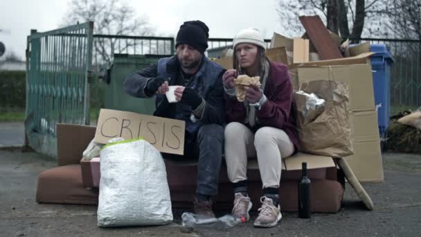两个衣衫褴褛的流浪汉 一个男人和一个女人 坐在一堆垃圾旁边 手里拿着一张手写的Crisis海报 吃着什么东西 — 图库视频影像