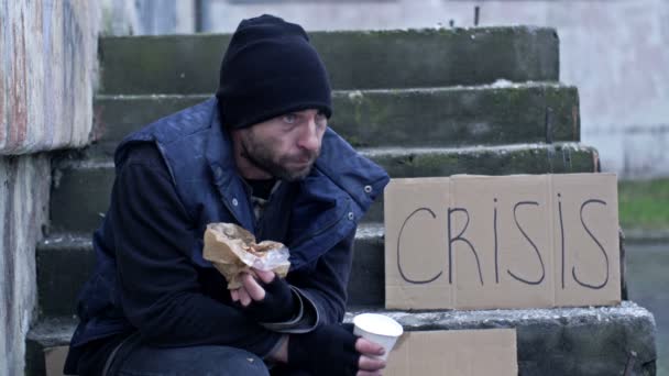 冷酷和衣衫褴褛的男人坐在外面的台阶上 吃着什么东西 旁边是一张手写的Crisis海报 — 图库视频影像