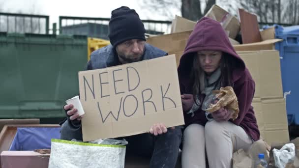 汚い服を着た2人のホームレスの人々 男性と女性は 手書きのNeed Workポスターでゴミの山に座っています — ストック動画