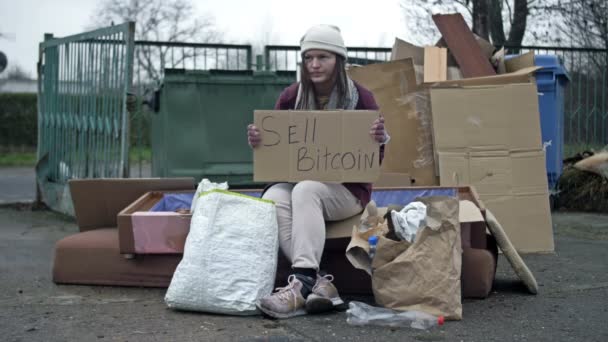 冷たい目立たない若い女性がゴミの山に座り 手書きのSell Bitcoinポスターを持っています 女は病気で咳をしている — ストック動画