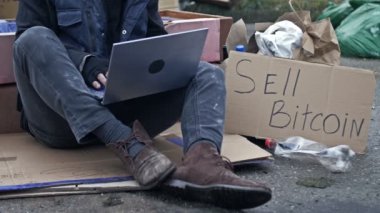 Bitcoin kazası yüzünden adam iflas etti. Kucağında dizüstü bilgisayarla soğukta titreyerek çöpün ortasında kartonda oturuyor. Yanında el yazısıyla satılan bir poster var. 4K.