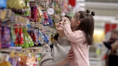 Güzel bir genç kadın ve küçük kızı süpermarkette şekerlerle rafların yanında. Çocuk parlak paketlerdeki şekerlemeleri seçiyor. 4K.