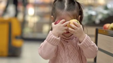Süpermarketteki küçük kız şakayla gözlerini elmalarla kapatıyor. HD.