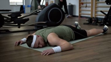 Fiziksel aktivitelerden yorulan genç şişman bir adam spor salonunda bir karemat üzerinde yatar. 4K