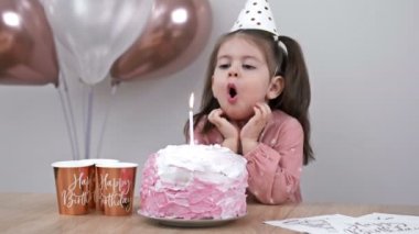 Little girl blows out candles on a birthday cake at home against a backdrop of balloons. Childs birthday. 4K.