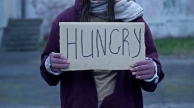 An unkempt-looking young woman sits with a begging cup and holds a handwritten HUNGRY poster. 4K.