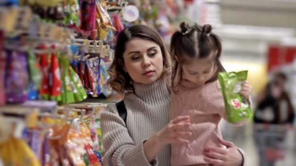 スーパーでお菓子と棚の近くの若い母親と子供 小さな女の子は明るい色のパッケージでキャンディを選択します — ストック動画