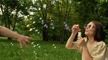 Sabun köpüğü üfleyen genç bir kadın. 6 yaşındaki kızı dans ediyor ve mutlu bir şekilde gökkuşağı kabarcıkları yakalıyor. Aile tatili. 4K.