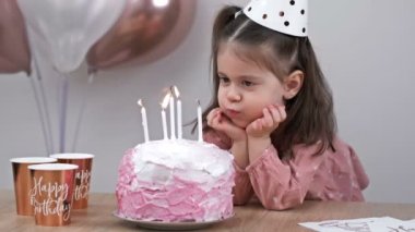 Little girl blows out candles on a birthday cake at home against a backdrop of balloons. Childs birthday. 4K.