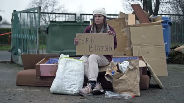 整頓されていない若い女性がゴミの山に座って 手書きのSell Bitcoinポスターを手に持っています 通行人が助けの代わりに不快な何かを言った停止しました — ストック動画