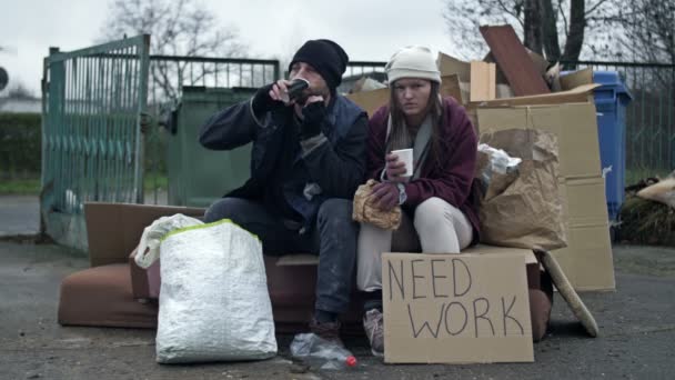 汚れた服を着た2人のホームレスの人々 男性と女性は 手書きのNeed Workポスターでゴミの山に座って何かを食べています 若い女性は病気で咳をしている — ストック動画