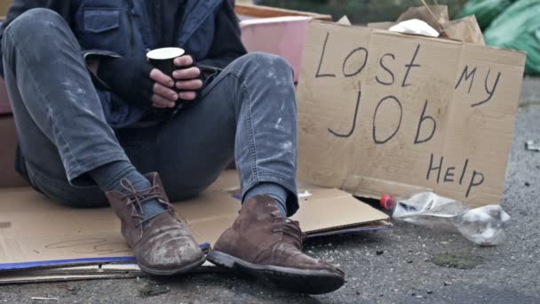 寒さに震えるホームレスの男がゴミの中に段ボールに座っている 彼の隣には手書きのLost Jobヘルプポスターがあります — ストック動画