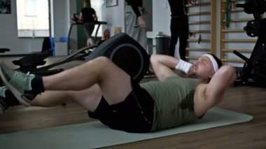 Kilolu bir adam spor salonunda paspasın üzerine uzanmış karın kaslarını özenle itiyor. 4K.
