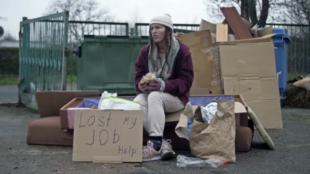 冷たい 汚れた若い女性がゴミの山に座って 貪欲に何かを食べる 近くに手書きのLast Jobヘルプポスターがあります — ストック動画