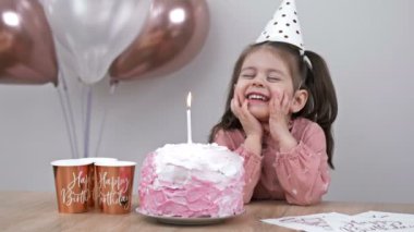 Little girl blows out candles on a birthday cake at home against a backdrop of balloons. Childs birthday. 4K.