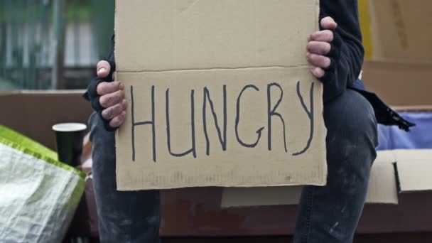 一个不整洁的男人坐在那里 端着一杯饮料 手里拿着一张手写的Hungry标语牌 — 图库视频影像