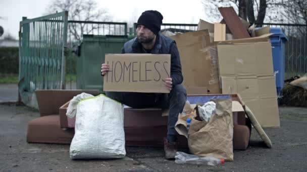 冷凍された片思いの青年はゴミの山に座り手書きのホームレスのポスターを持っている — ストック動画