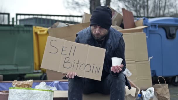 冷冰冰的 不修边幅的年轻人坐在一堆垃圾旁边 手里拿着一个乞讨杯 举着一张手写的 卖东西的双边投资条约 — 图库视频影像