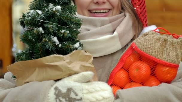 クリスマスフェアで美しい金髪の女性 女性は小さなクリスマスツリーと明るい色のタンジェリーンを保持しています メリークリスマス 4Kについて — ストック動画