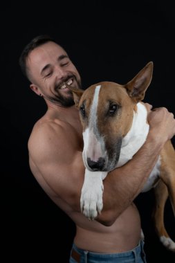 Siyah arka planda güçlü üstsüz bir adam köpeğine sarılıyor.
