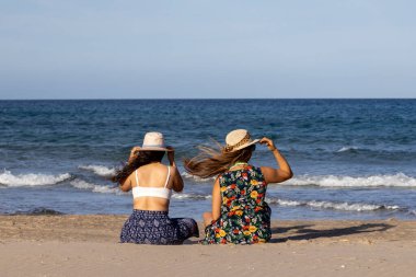 Arkadan kumsalda oturan şapkalı iki kız.