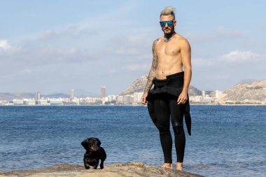 Bir adam köpeğiyle dalgıç kıyafetiyle denizde poz veriyor..