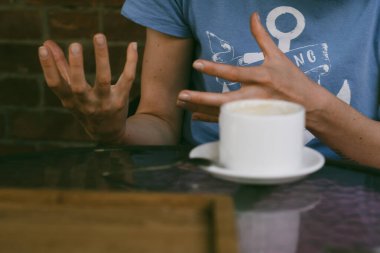 Bir kız bir kafede oturmuş sıcak bir yudum içmek için fron beyaz seramik bardak aynı anda hem konuşuyor hem de el kol hareketi yapıyor.