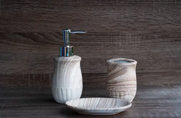a light gray liquid soap container with a silver dispenser, a cup for toothbrush and soap dish  on wooden background. Useful hygiene products and personal are. Place for texting or logo