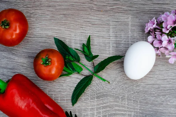 still life of fresh two tomatoes, one red pepper, one egg, mint for the salad, a bunch of violets on a wooden background, close up. Top view. Place for text or logo. Healthy eating concept.