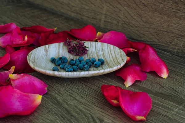 a soap dish with petals of pink rose, blue bilberry, purple violets on wooden background. Useful hygiene products and personal care. Place for texting or logo