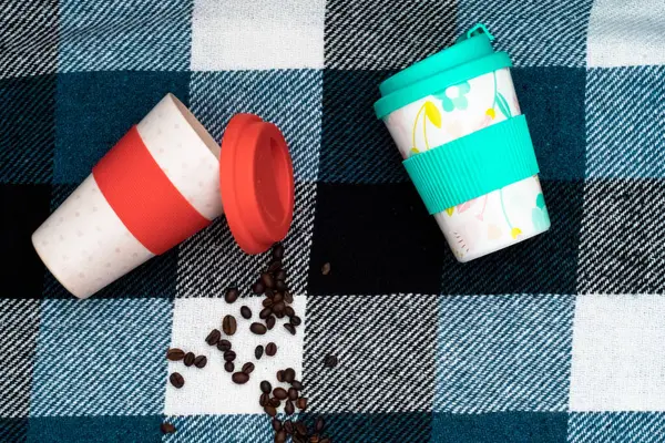 lying red and white cup with red lid and blue and white cup with blue lid, spilled coffee beans on plaid in a large blue and white check. Top view