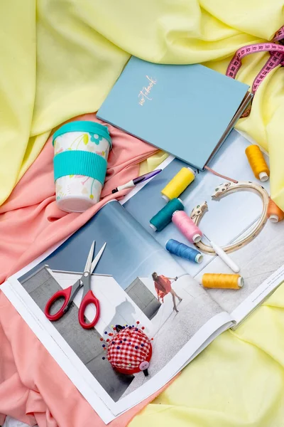 a closeup shot of red scissors, measuring tapes and blue and white plastic cup. threads, blue notebook on yellow and pink fabric