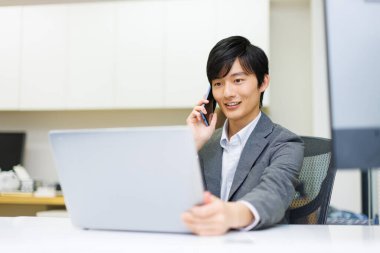 Genç Japon işadamı ofiste çalışırken telefonla konuşuyor..