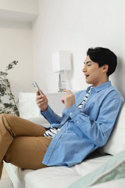 Genç Asyalı adam koltukta otururken cep telefonu kullanıyor.